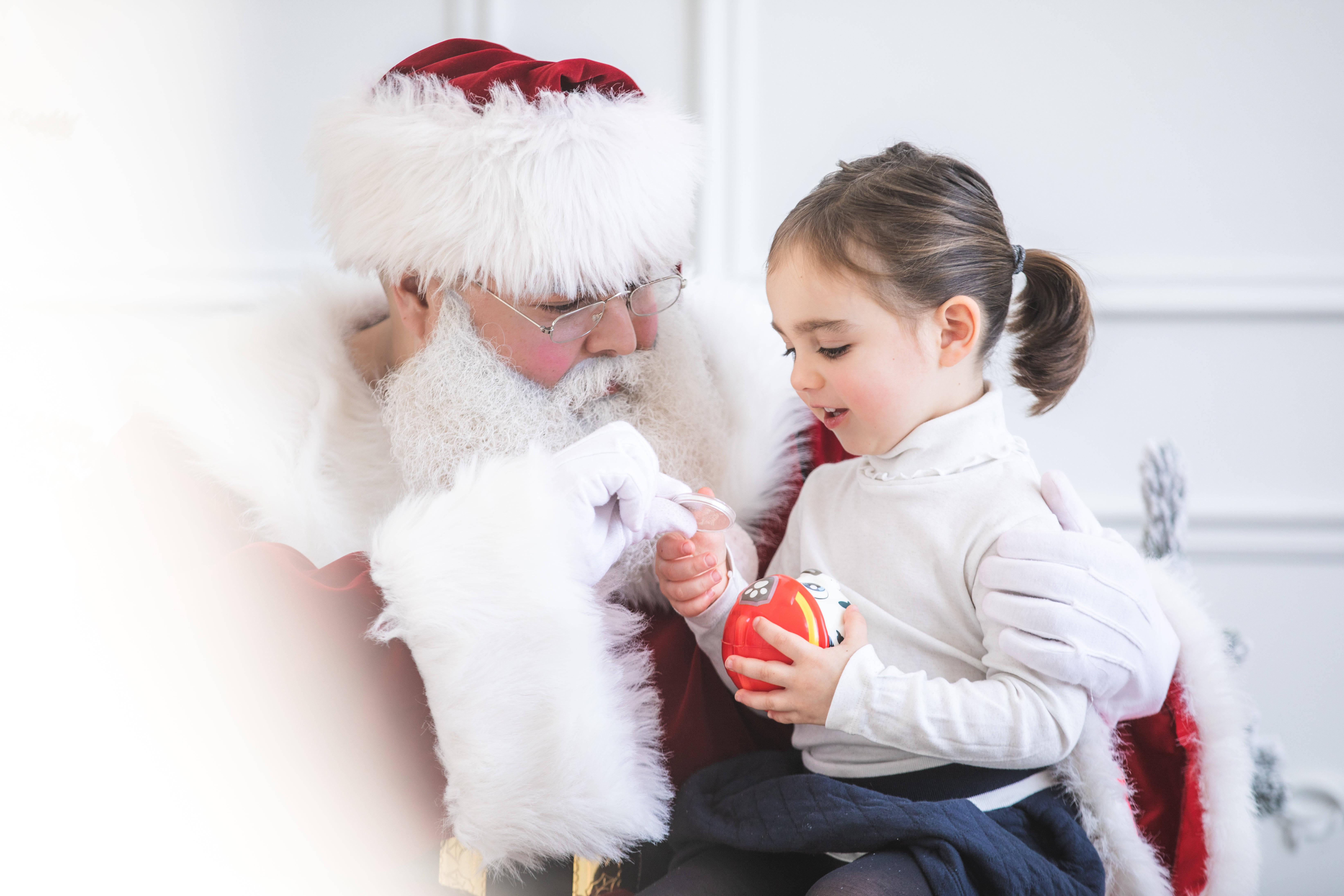 Santa Claus 桌布 壁紙 — 聖誕老人輕輕握住一個孩子的手，她正在明亮的白色房間裡檢查一個紅色玩具。