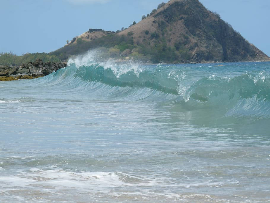 聖露西亞海岸壯麗海浪 壁紙 — 一道強勁的藍綠色海浪在岩石海岸附近翻捲並破碎，背景是崎嶇的山丘和晴朗的藍天。