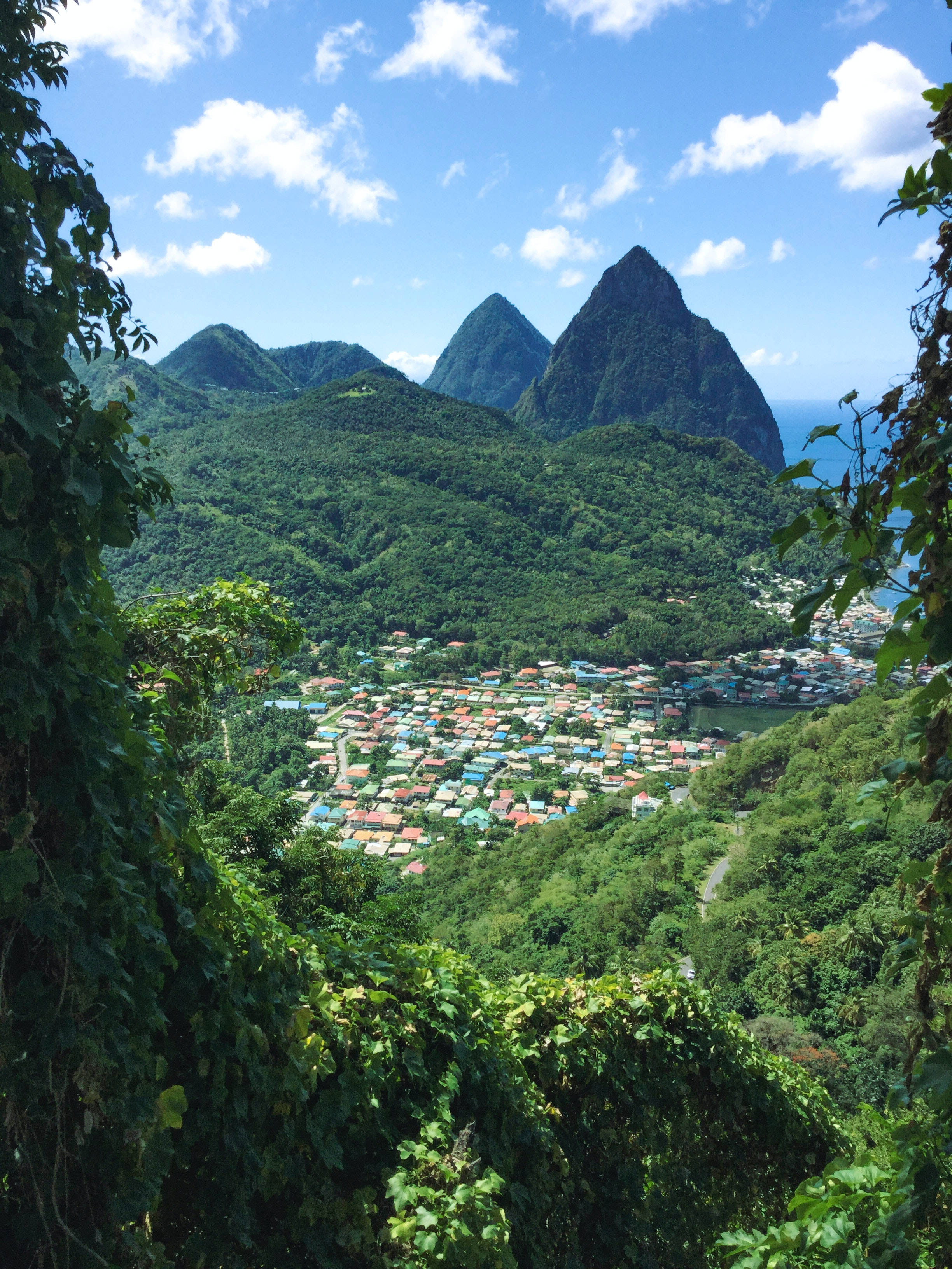 聖露西亞自然風景 壁紙 — 一個充滿活力的村莊坐落在鬱鬱蔥蔥的綠色山谷中，下方是雄偉的火山峰，上方是明亮的藍天。