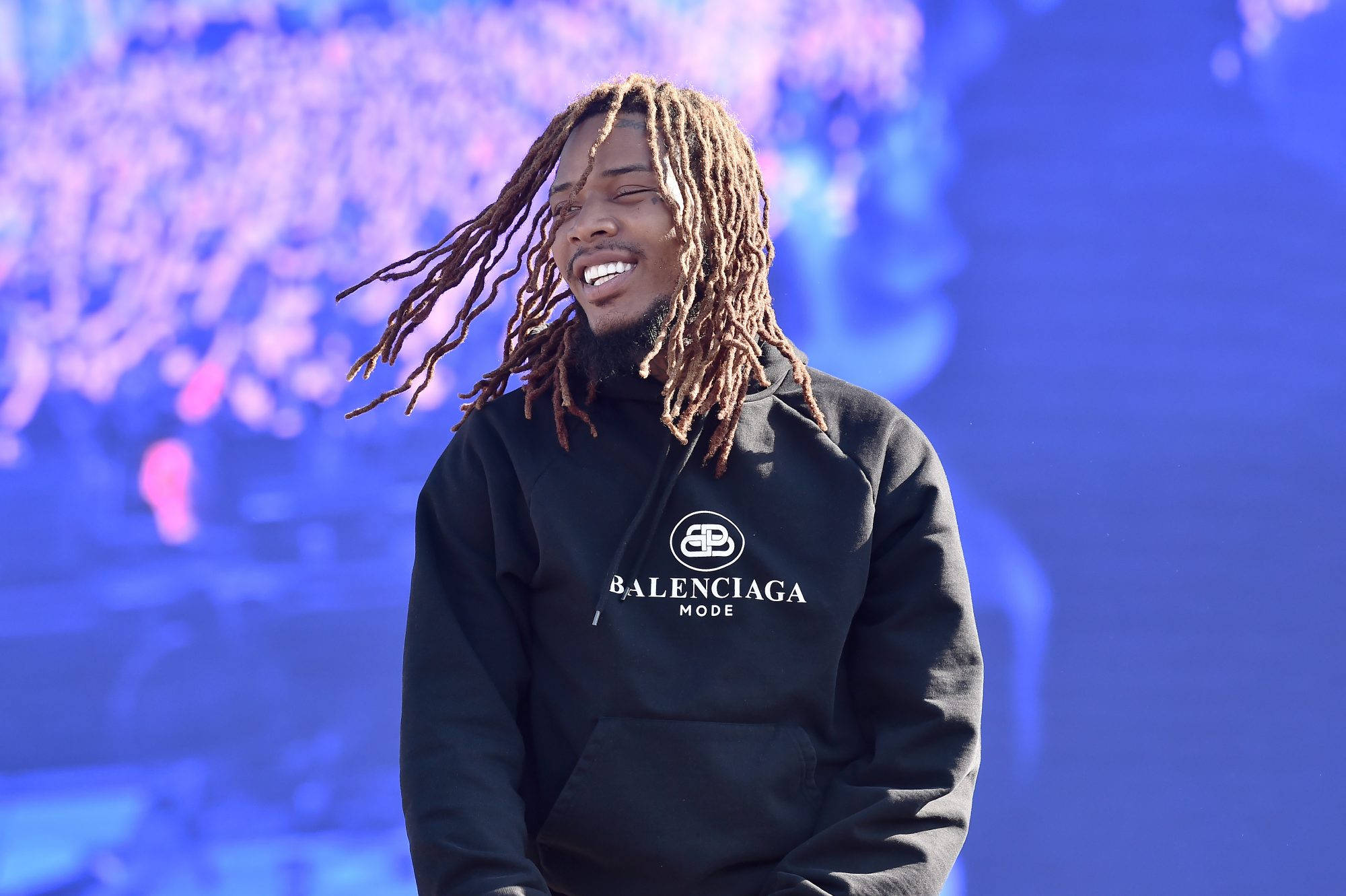 聚焦Fetty Wap 壁紙 — 一位微笑的長髮辮男士穿著黑色Balenciaga連帽衫，背景是鮮豔的藍色和紫色模糊效果。