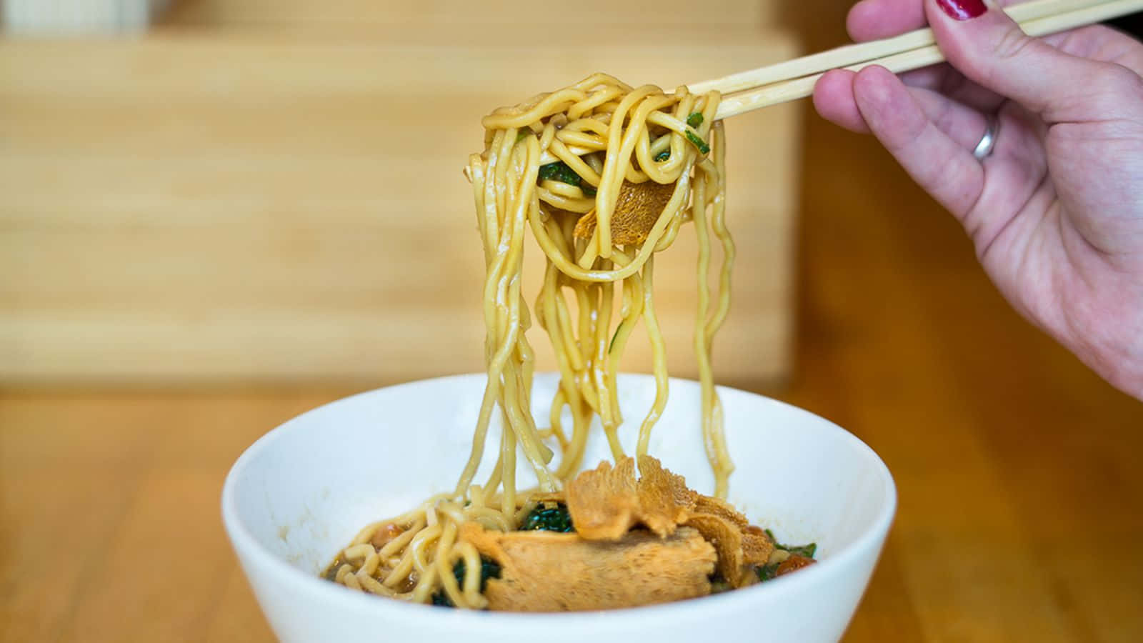 聚焦乾麵條的特寫照片 壁紙 — 一隻手用筷子從裝滿濃郁湯頭和豆腐的白色碗中夾起冒著熱氣的麵條。