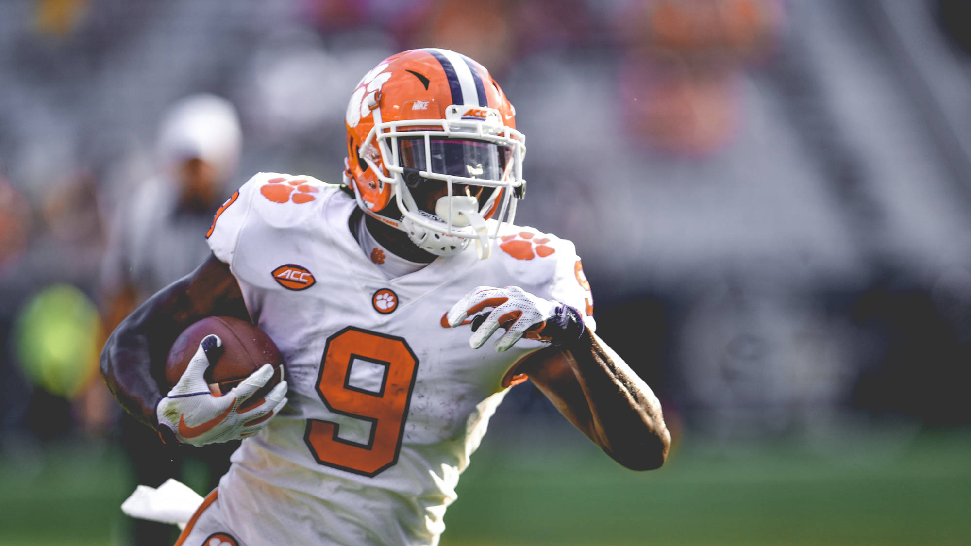 聚焦拍攝 Travis Etienne 場上比賽 壁紙 — 一名身穿白色和橙色裝備的克萊姆森足球員在日間比賽中持球衝刺。