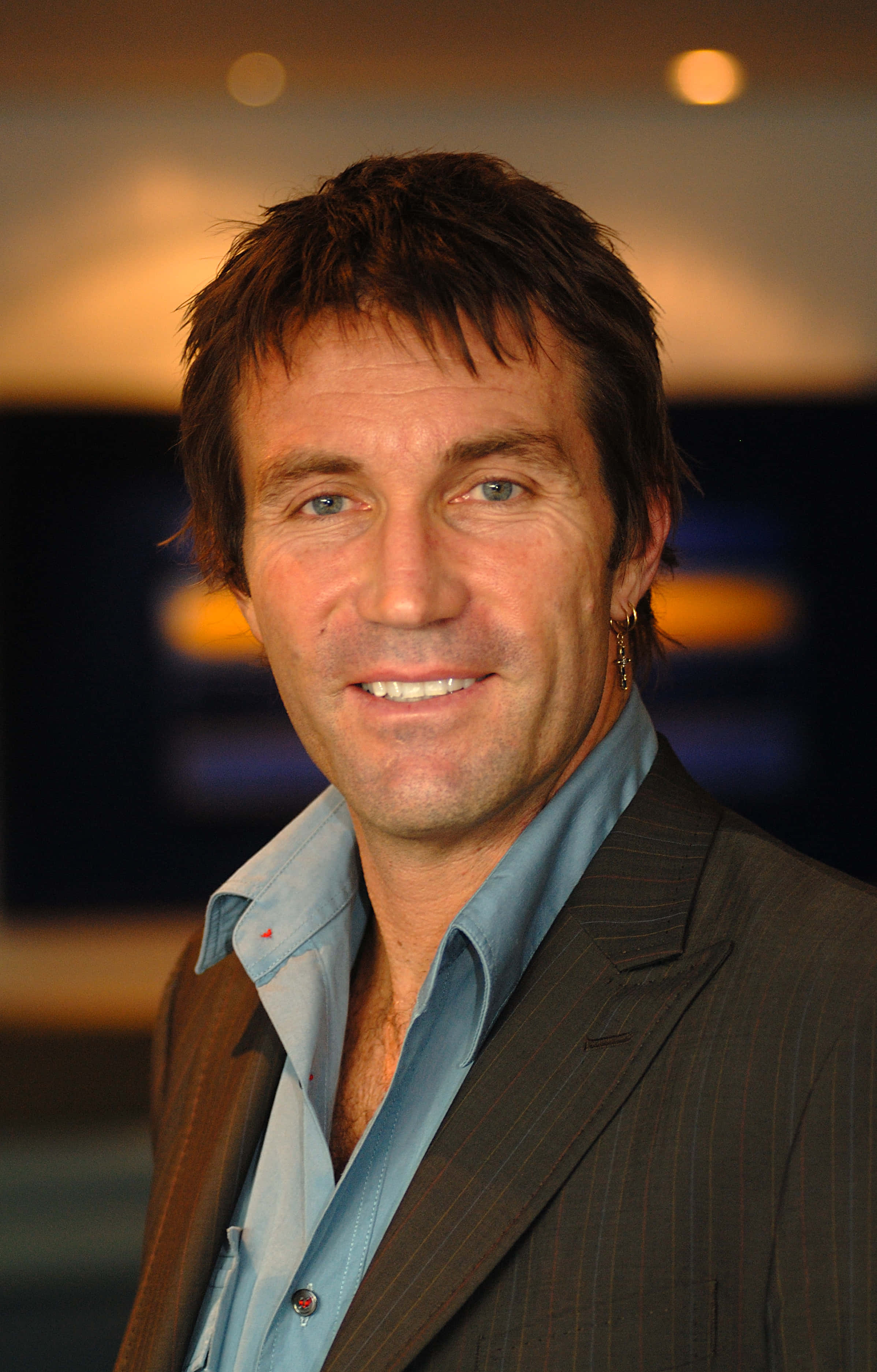 聚焦照片：澳洲網球冠軍 Pat Cash 壁紙 — 一位短黑髮藍眼的微笑男士，身穿細條紋西裝，內搭淺藍色襯衫，背景是溫暖燈光的模糊效果。