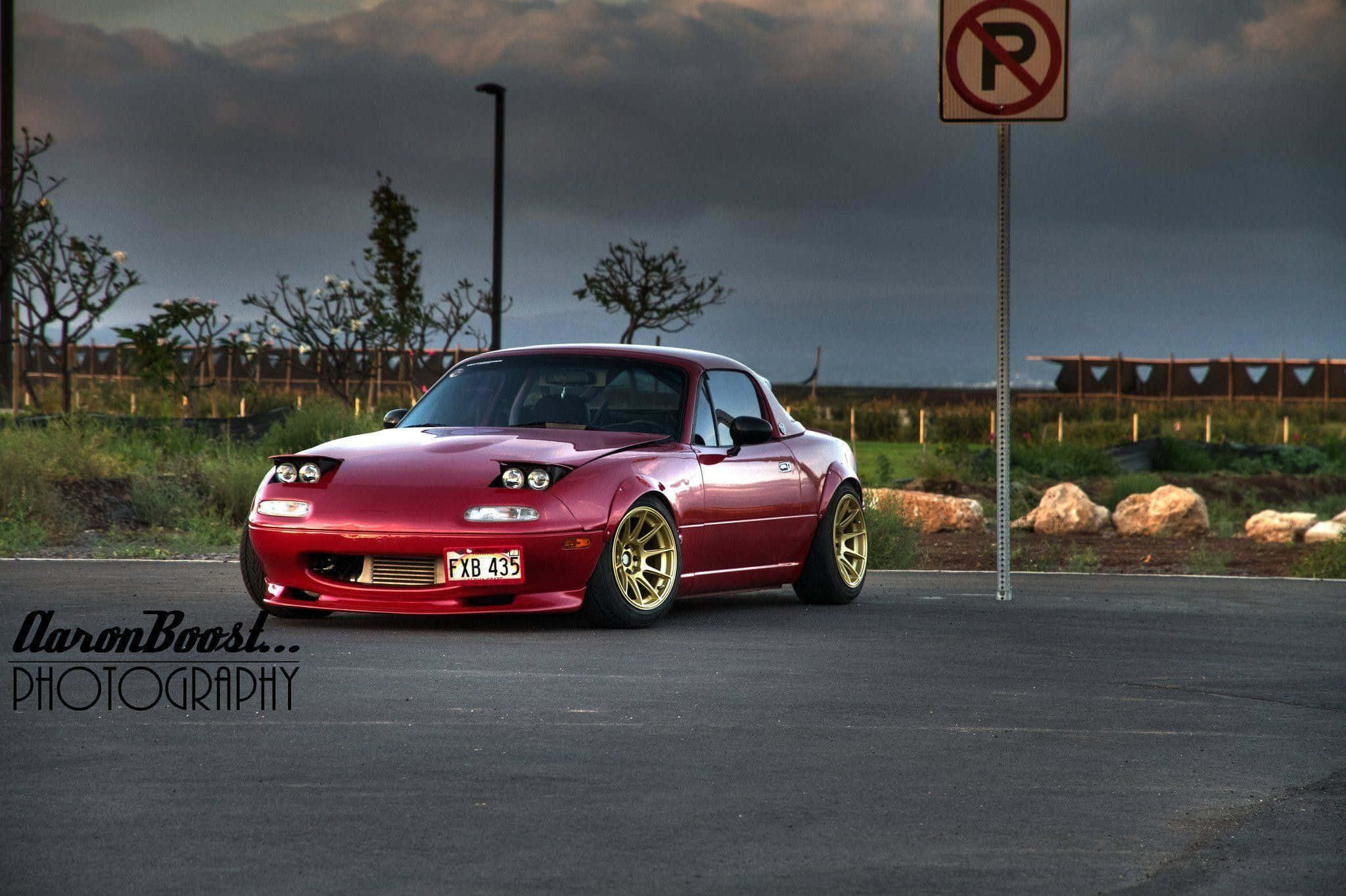 聚焦馬自達MX-5 Miata 壁紙 — 一架光澤紅色Mazda Miata配金色輪圈，停泊在空地上，背景是風暴前夕的暮色天空。