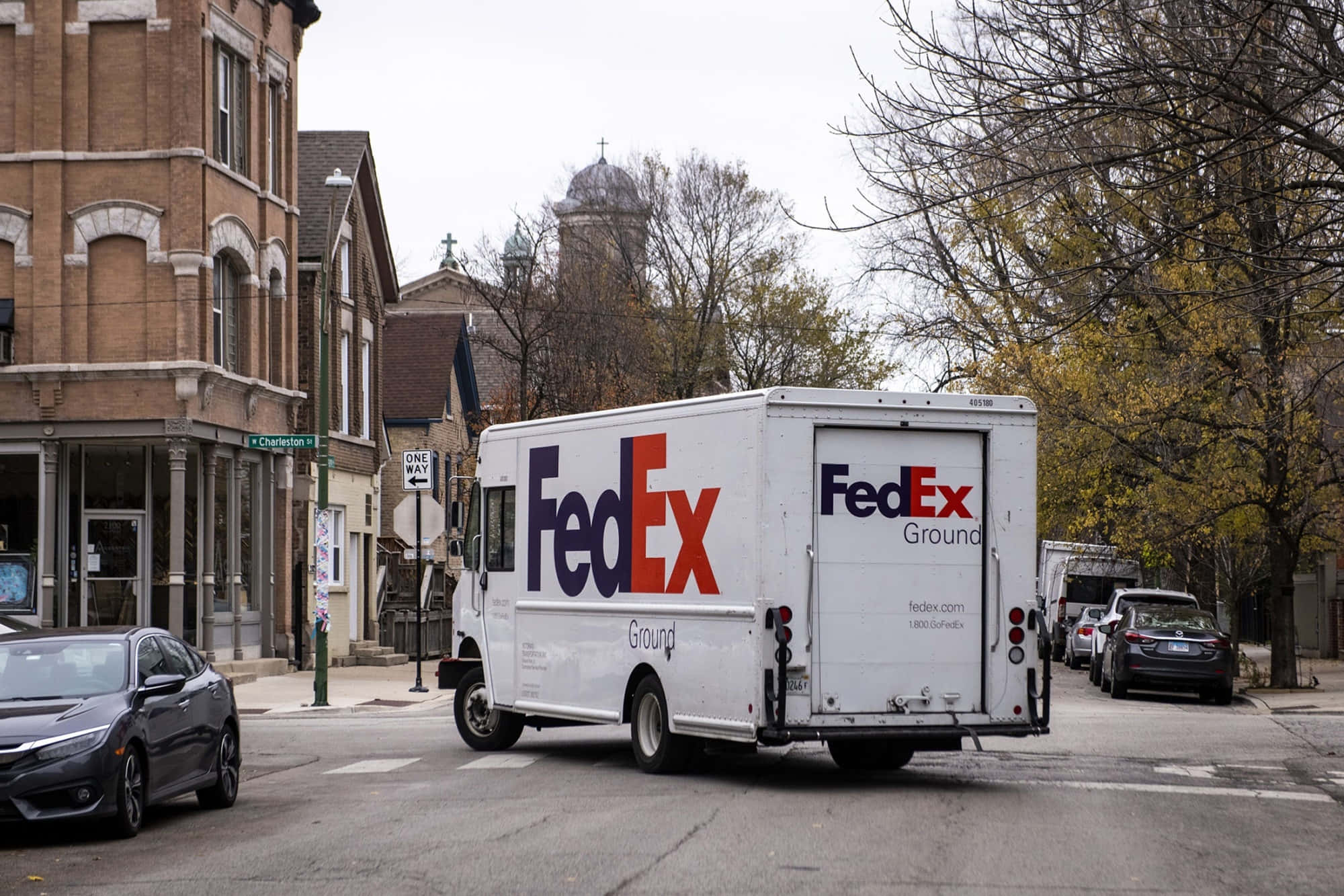 白色FedEx Ground送貨車在城市街道上，旁邊有磚砌建築和秋天的樹木。