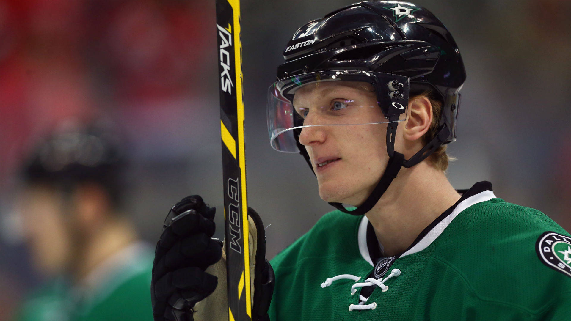 職業NHL球員約翰·克林格伯格賽場表現 壁紙 — 一位專注的曲棍球運動員身穿綠色球衣，在明亮的場館燈光下於激烈的比賽中握著球棍。