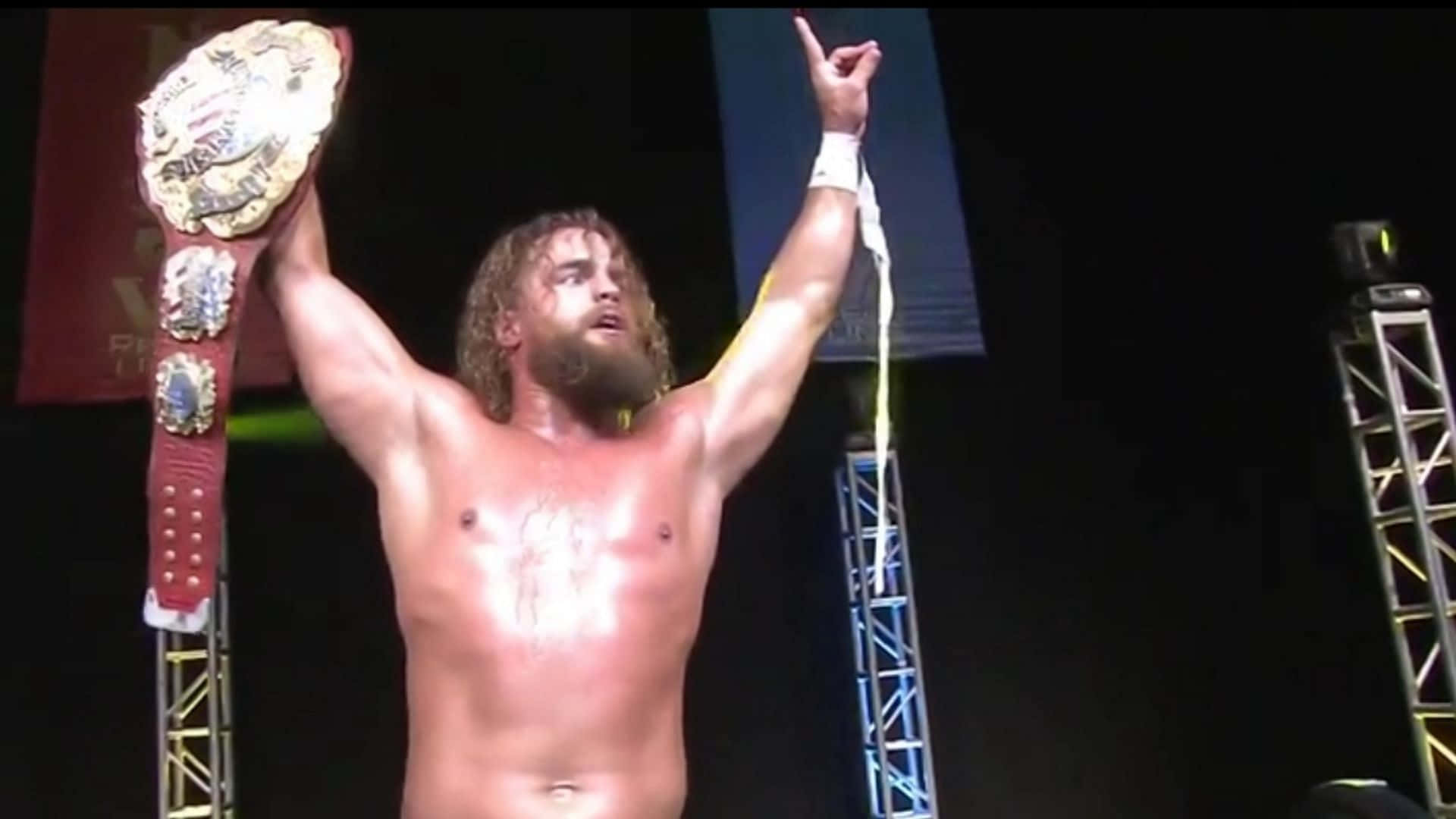 職業摔角手Juice Robinson 壁紙 — 一位汗流浹背、留鬍子的摔角手，在明亮的舞台燈光下，一手高舉冠軍腰帶，另一手舉起勝利手勢。