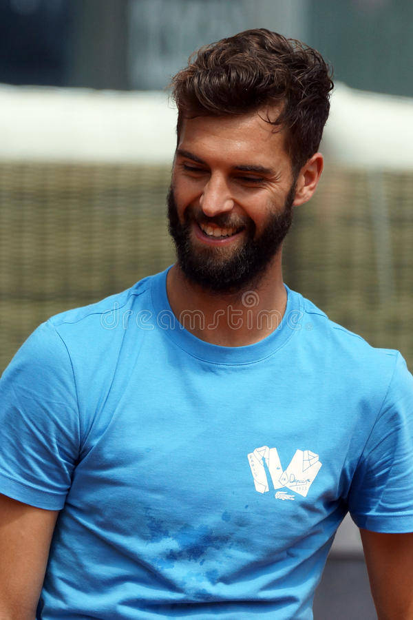 職業網球選手 Benoit Paire 微笑側影 壁紙 — 一位留鬍鬚的男子身穿藍色T恤，在模糊的城市背景下，眼睛因真摯的喜悅而瞇起，溫暖地微笑。