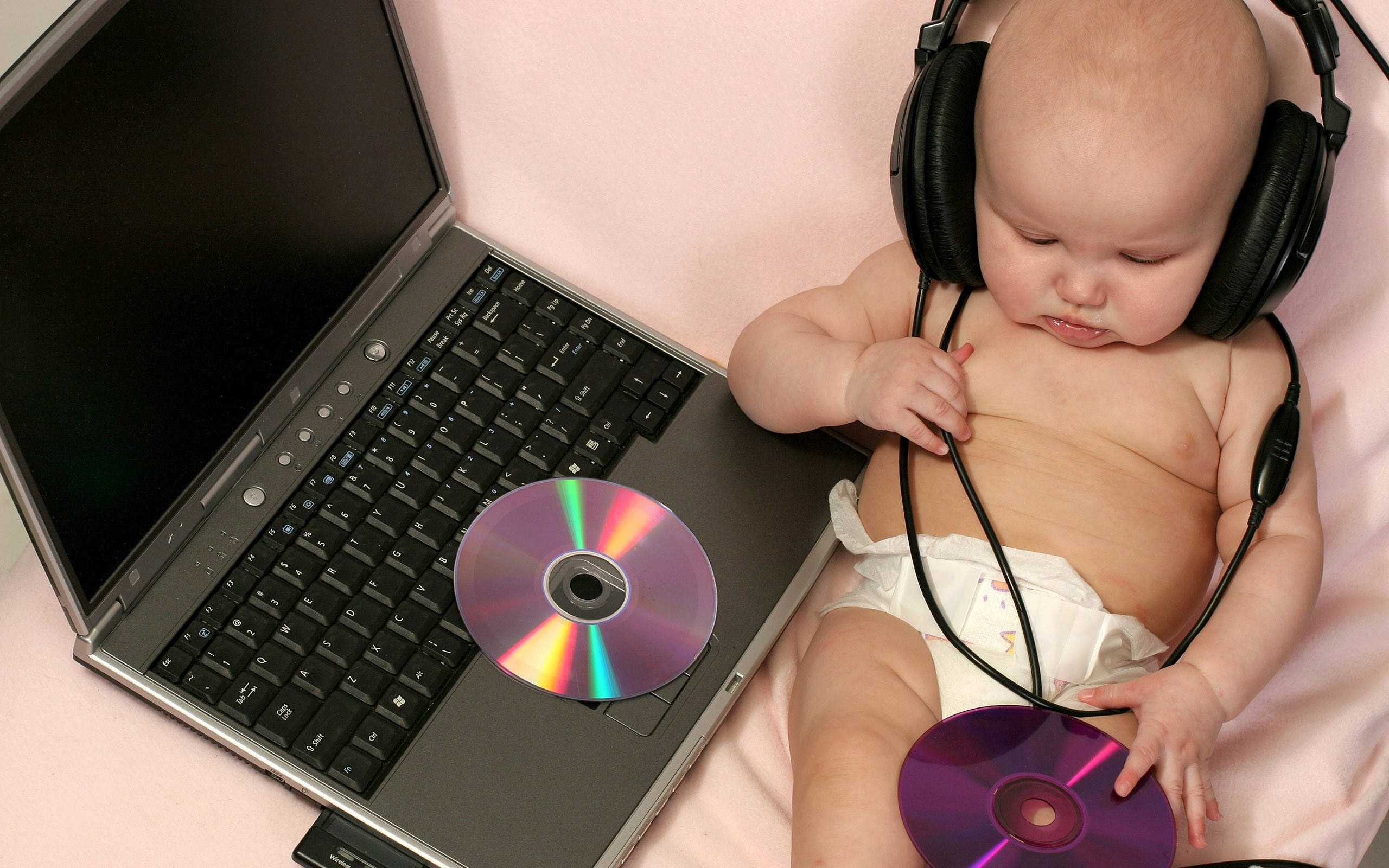 聽音樂的搞笑嬰兒 壁紙 — 一個穿著尿布的嬰兒戴著特大耳機，在粉紅色毯子上打開的筆記本電腦旁邊拿著CD，安詳地睡著。