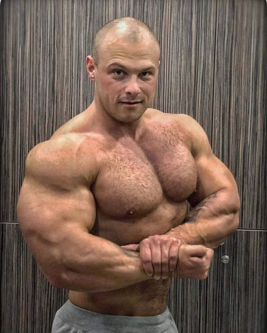 肌肉男擺姿圖片 壁紙 — 一名赤裸上身的肌肉男，在垂直的木鑲板牆前用力彎曲手臂，神情專注。