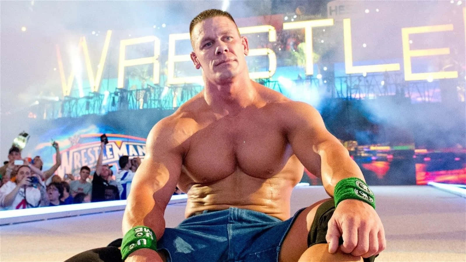 John Cena 桌布 壁紙 — 肌肉發達的摔角手約翰·辛納在明亮的場館燈光下自信地坐在擂台上，背後閃爍著「WRESTLEMANIA」字樣。