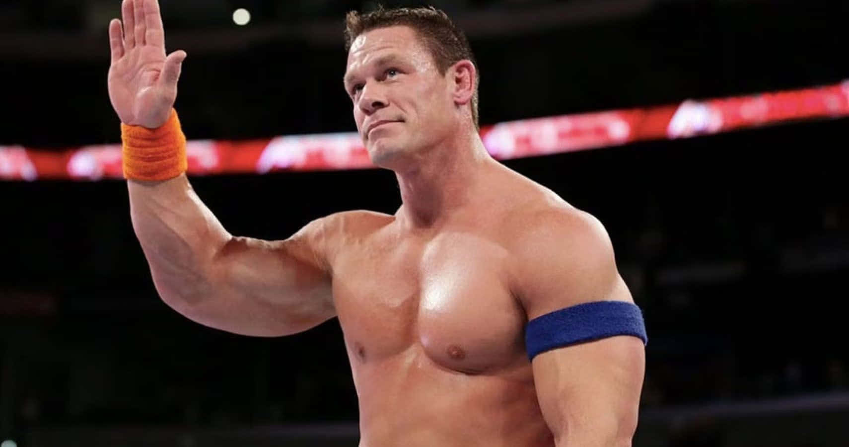 John Cena 桌布 壁紙 — 肌肉發達的摔角手約翰·辛納在燈光明亮、背景有紅光的擂台上向粉絲揮手。