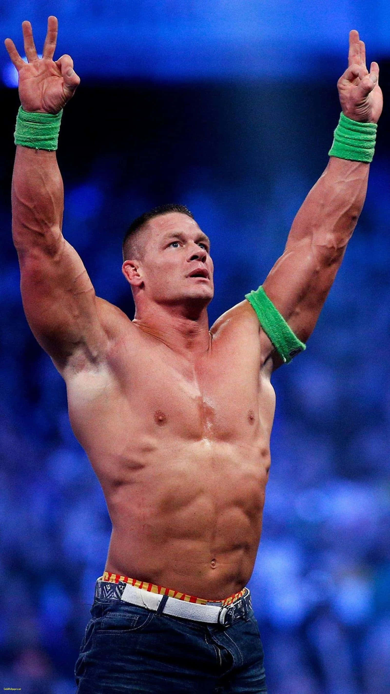 John Cena 桌布 壁紙 — 肌肉發達的摔角手約翰·辛納戴着綠色腕帶，在模糊的藍色競技場人群前高舉雙臂慶祝勝利。