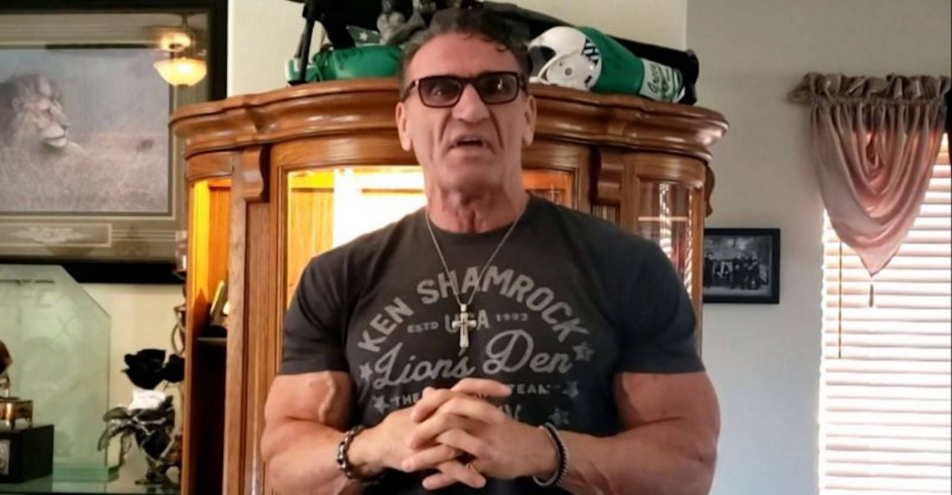 肌肉發達的男人戴著太陽眼鏡和 Ken Shamrock T恤，站在一個有獎盃的房間裡。