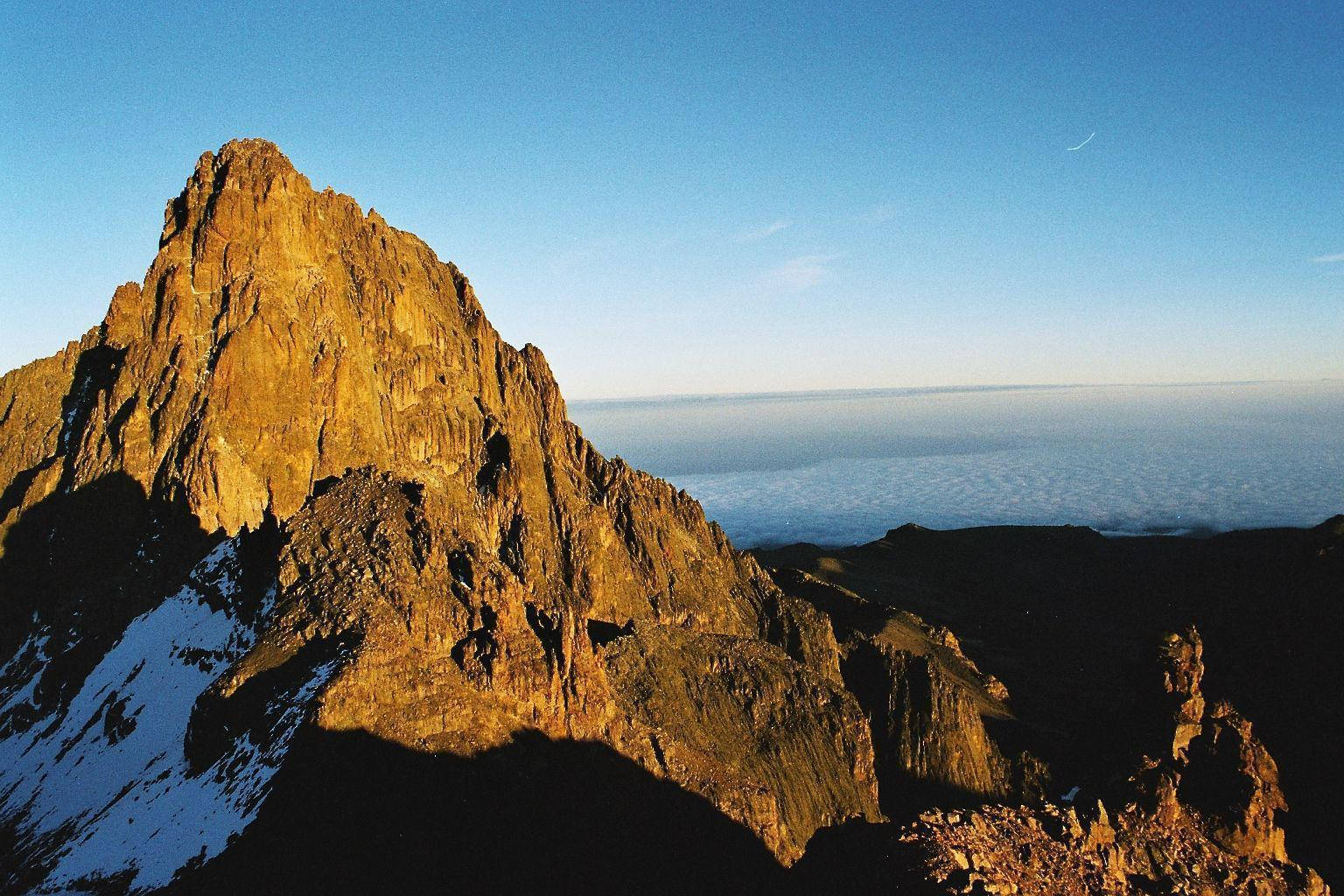 肯亞非洲岩石山脈 壁紙 — 一個崎嶇的山峰沐浴在金黃色的日出光芒中，聳立在雲海之上，陰影籠罩的山坡上還掛著積雪。