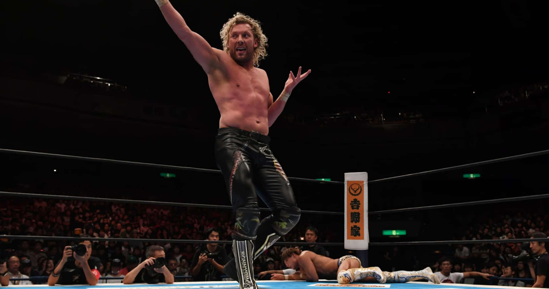 肯尼·歐米加於King of Pro-Wresting賽事中揮手致意 壁紙 — 一位赤膊、捲髮的摔角手在擂台上勝利地跳躍，而他的對手則被打敗躺在下方。