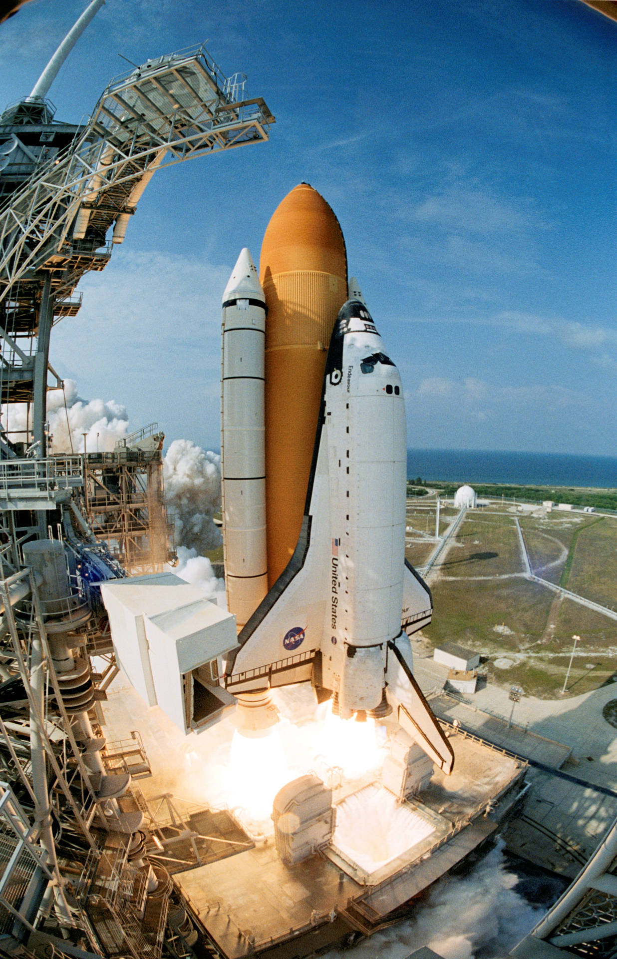 肯尼迪航天中心 STS-111 航天飛機發射 壁紙 — 美國太空總署的太空穿梭機在發射台上點燃引擎，噴出熾熱的廢氣和滾滾濃煙，映襯著清澈的藍天。