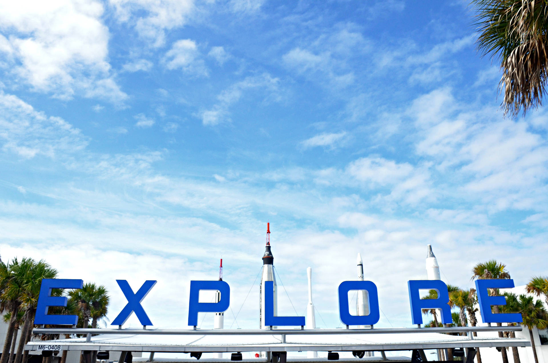 藍天下的藍色「EXPLORE」標誌，伴隨著棕櫚樹和火箭模型。 — Nasa GIF 壁紙