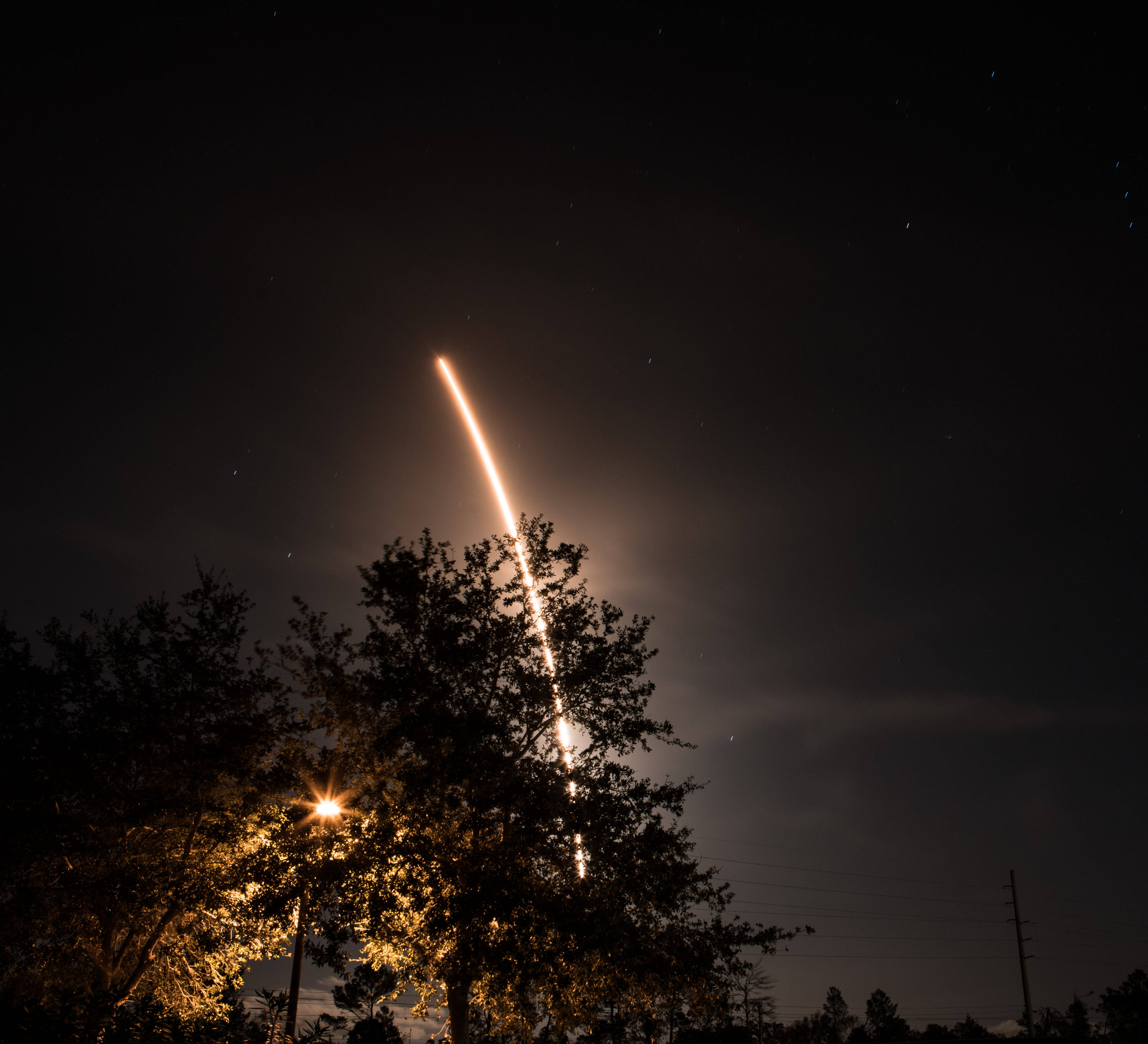 肯尼迪航天中心太空船發射 壁紙 — 一枚明亮的火箭在繁星點點的夜空中上升，留下發光的軌跡，而剪影般的樹木和街燈框住了下方的場景。