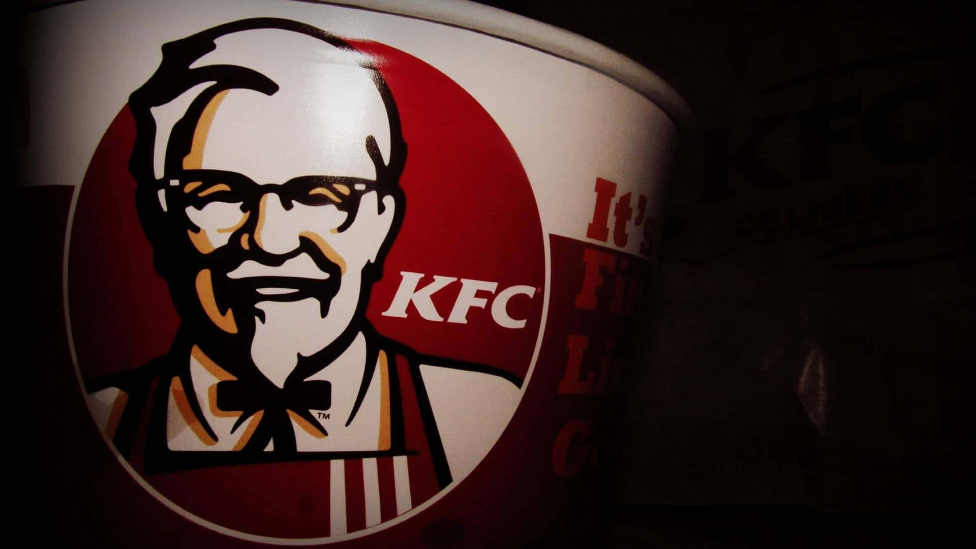 肯德基經典炸雞——您最愛的快餐目的地！ 壁紙 — KFC 桶的特寫，在戲劇性的低光照明下展示山德士上校的標誌性標誌。