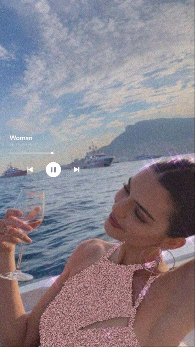 肯豆「Woman」 壁紙 — 一位身穿閃亮粉紅色上衣的女性在雲層密布的天空下，於一艘有遊艇和山脈的船上啜飲葡萄酒。