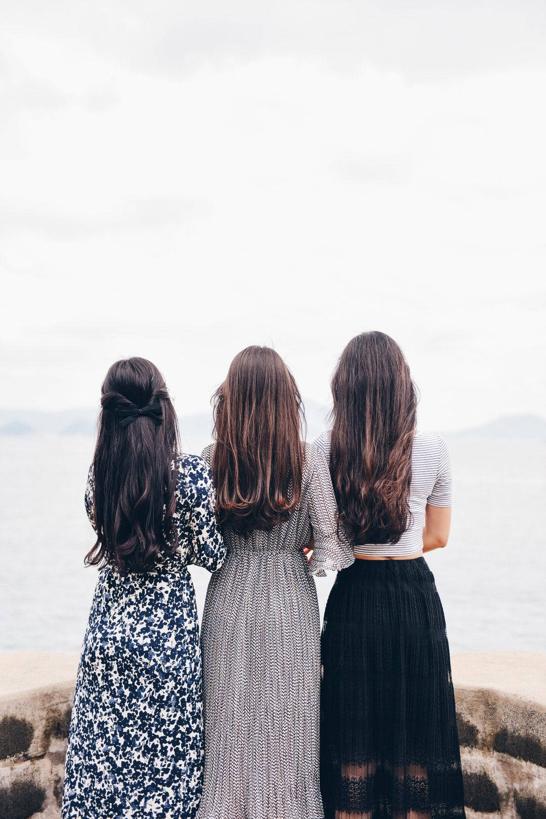 背對觀景的三位女孩 壁紙 — 三位長髮女性背對背站在岩石懸崖上，俯瞰著陰天下的平靜大海。