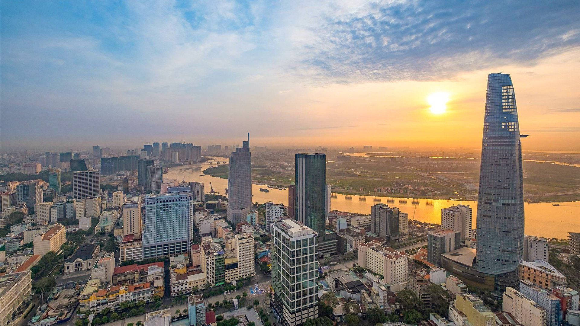 胡志明市日出 壁紙 — 夕陽將廣闊的現代城市天際線沐浴在金色光芒中，摩天大樓在寬闊的河流旁聳立。