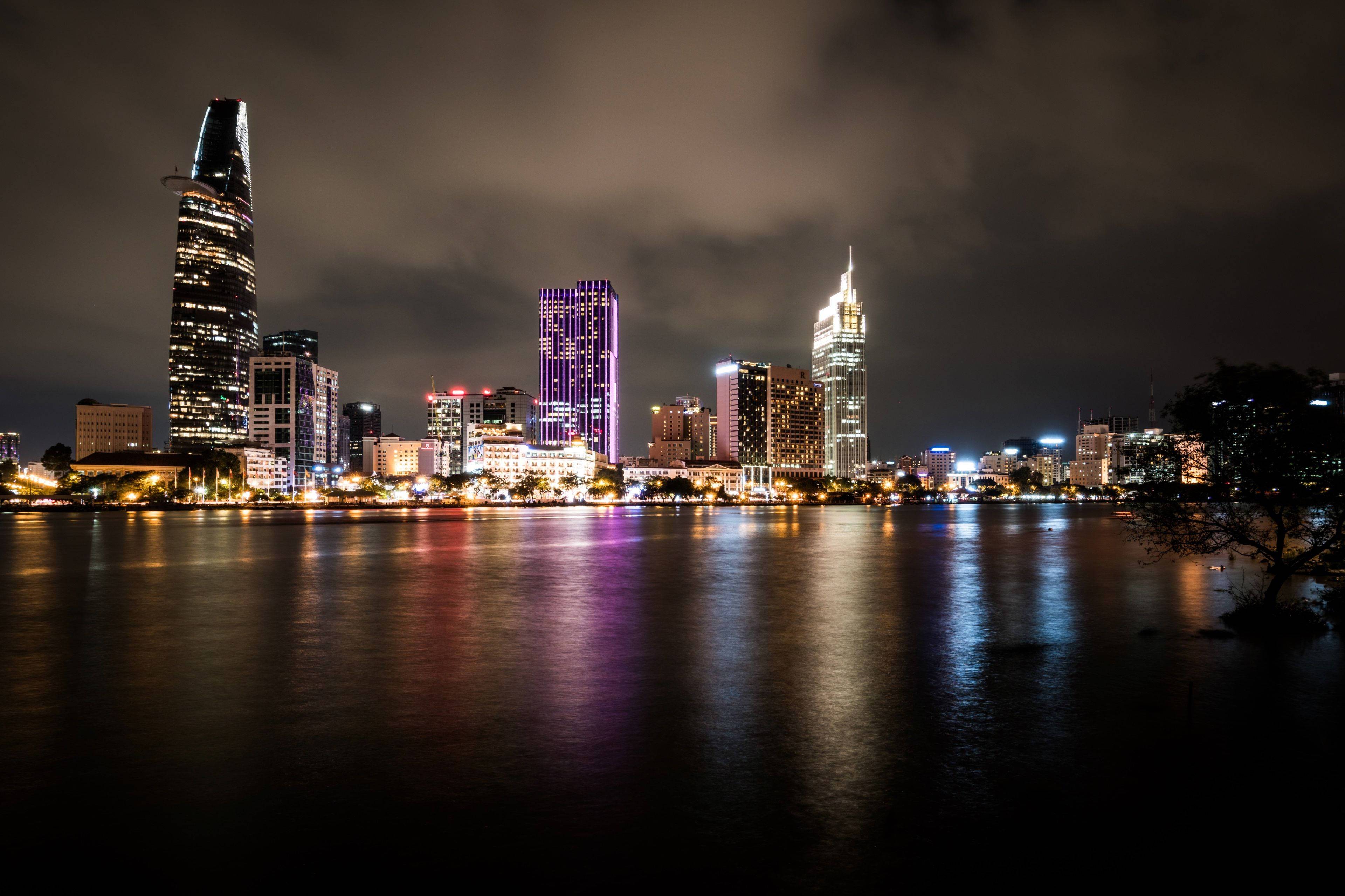 胡志明市暗色水域 壁紙 — 充滿活力的夜景城市景觀，在陰天之下，燈火通明的摩天大樓倒映在平靜的水面上。