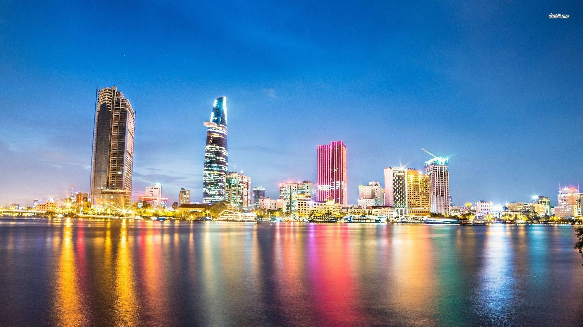 胡志明市林立建築 壁紙 — 黃昏時分充滿活力的城市天際線，色彩繽紛的摩天大樓在深藍色天空下平靜的水面上倒映。