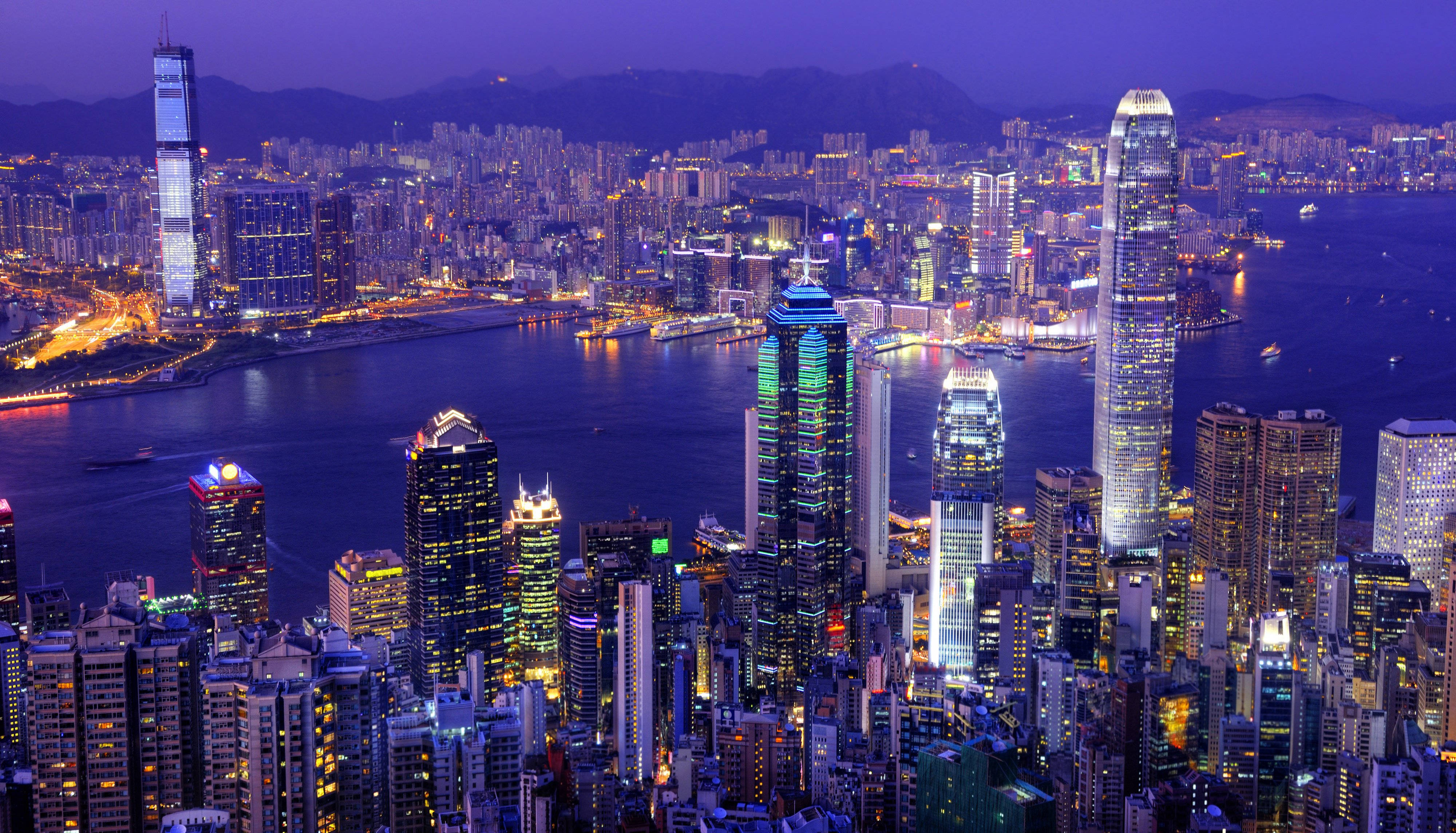 胡志明市河畔城市 壁紙 — 香港天際線在暮色中的迷人景觀，閃爍的摩天大樓和充滿活力的港口倒映著城市的燈光。