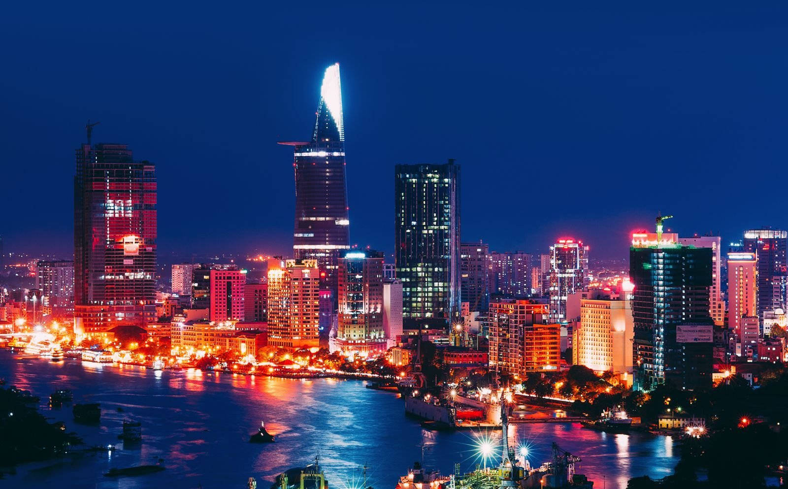 胡志明市絢麗燈光 壁紙 — 充滿活力的夜景，在深藍色天空下，發光的摩天大樓倒映在閃爍的河流上。