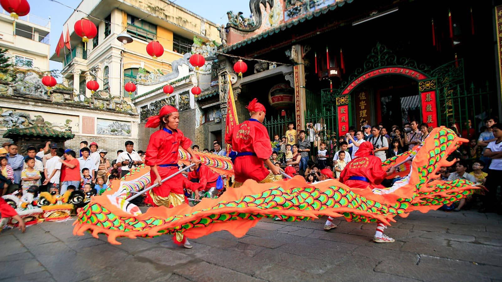 胡志明市舞龍表演 壁紙 — 鮮豔的橙色舞龍者穿著紅色服裝，在裝飾著紅色燈籠的寺廟前進行傳統舞蹈，人群圍觀。