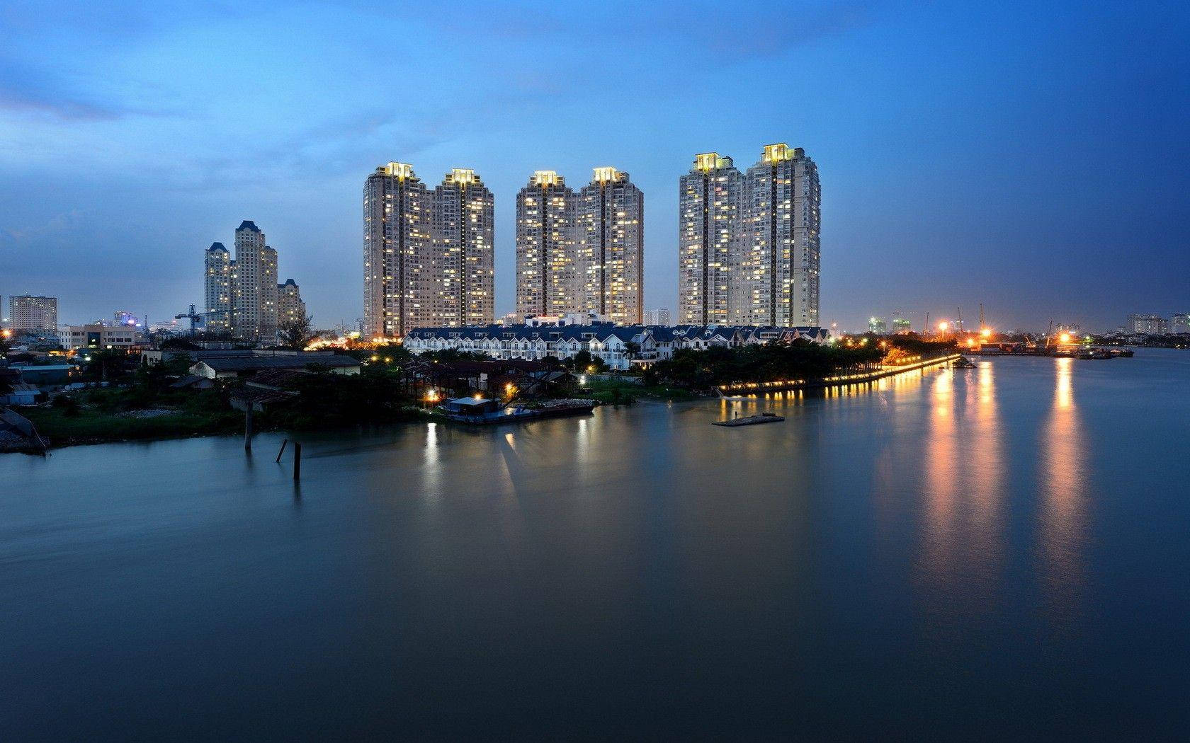 胡志明市西貢珍珠塔 壁紙 — 高聳的燈火通明摩天大樓在暮色中倒映在平靜的水面上，遠處有橋樑閃爍著光芒。