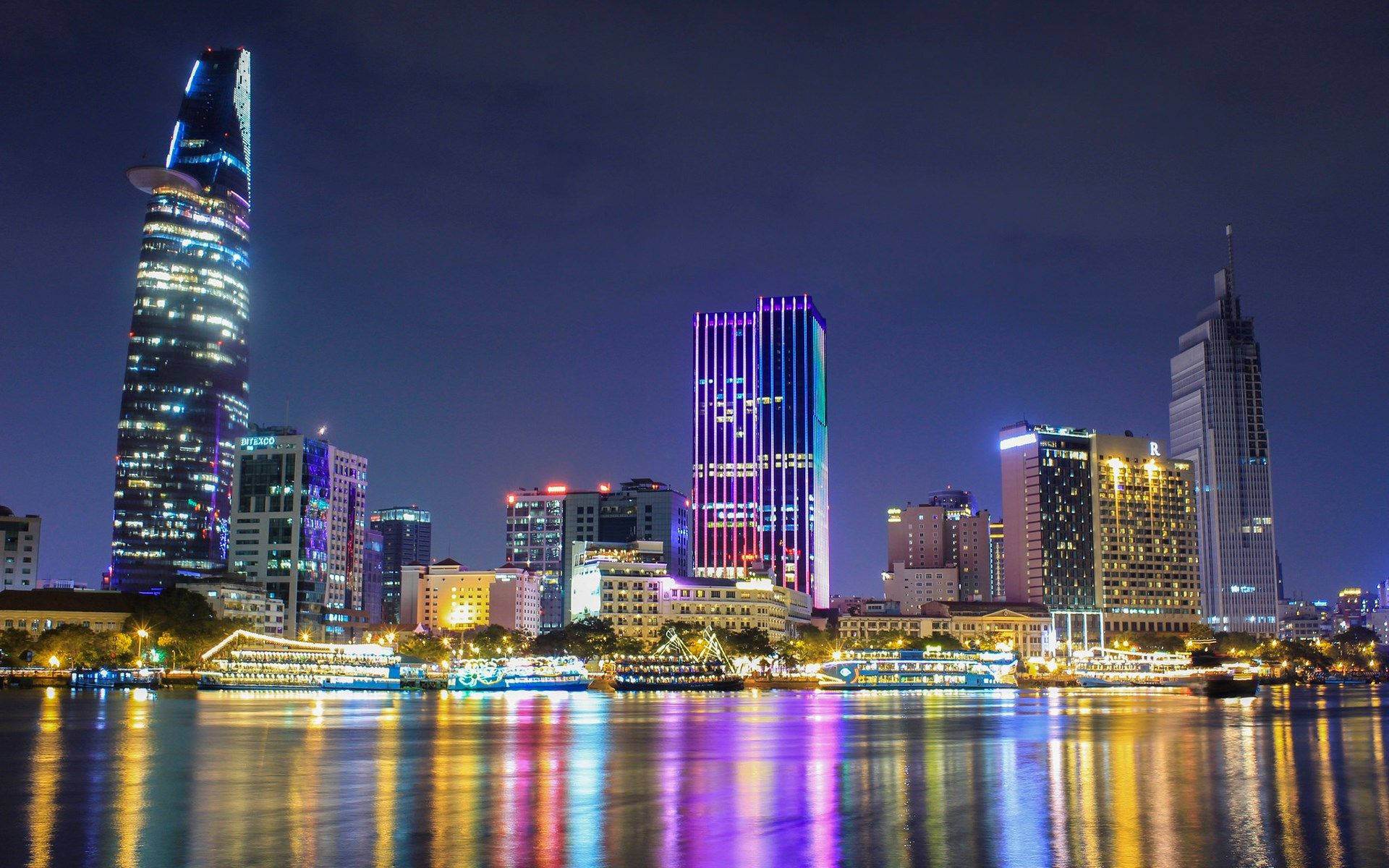 胡志明市迷人夜景 壁紙 — 夜晚充滿活力的城市天際線，燈火通明的摩天大樓倒映在下方平靜的水面上。