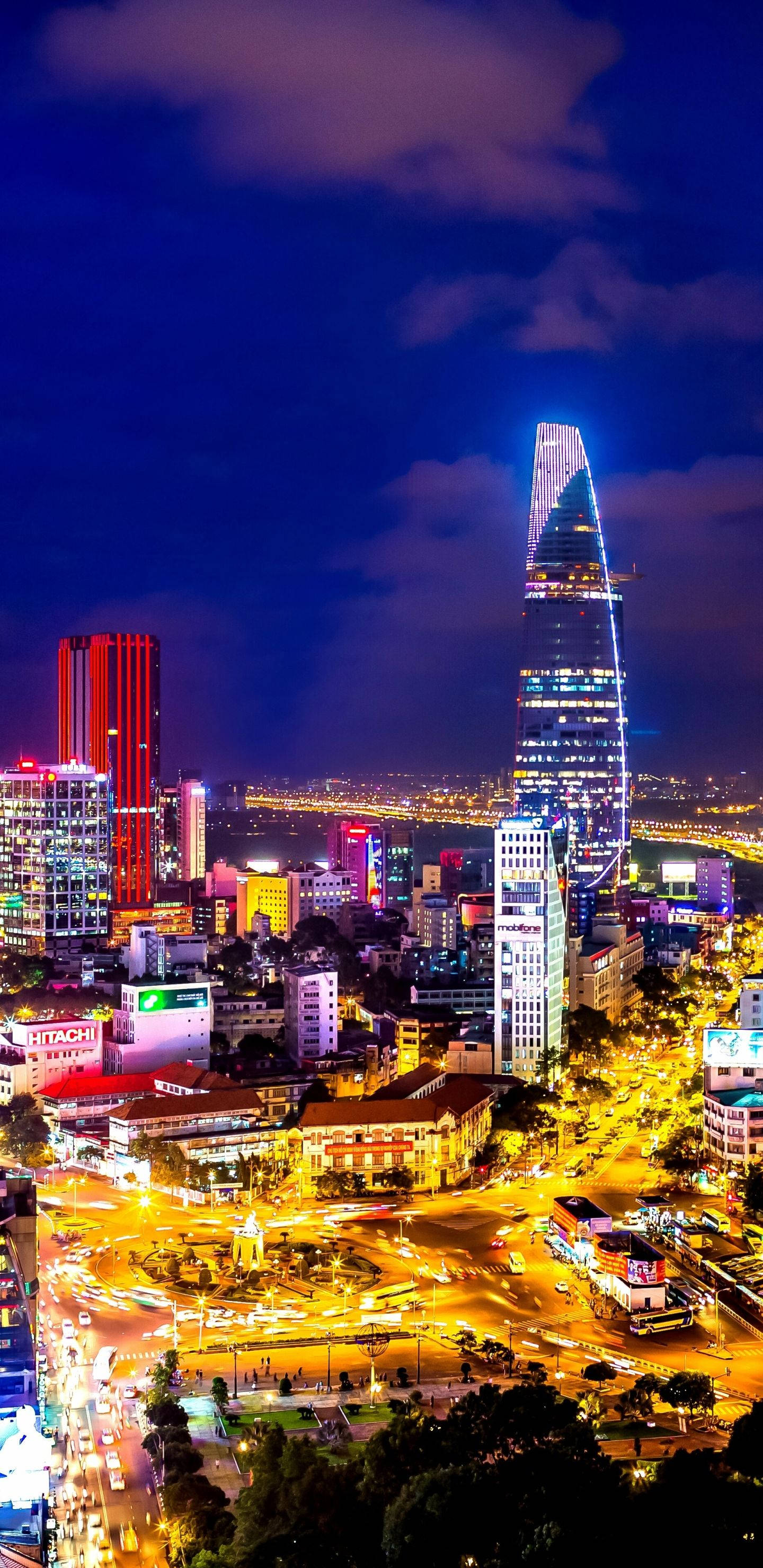 胡志明市金融大廈 壁紙 — 充滿活力的夜景城市景觀，一座高聳的摩天大樓在深紫色天空和下方繁忙街道的映襯下發出藍色光芒。