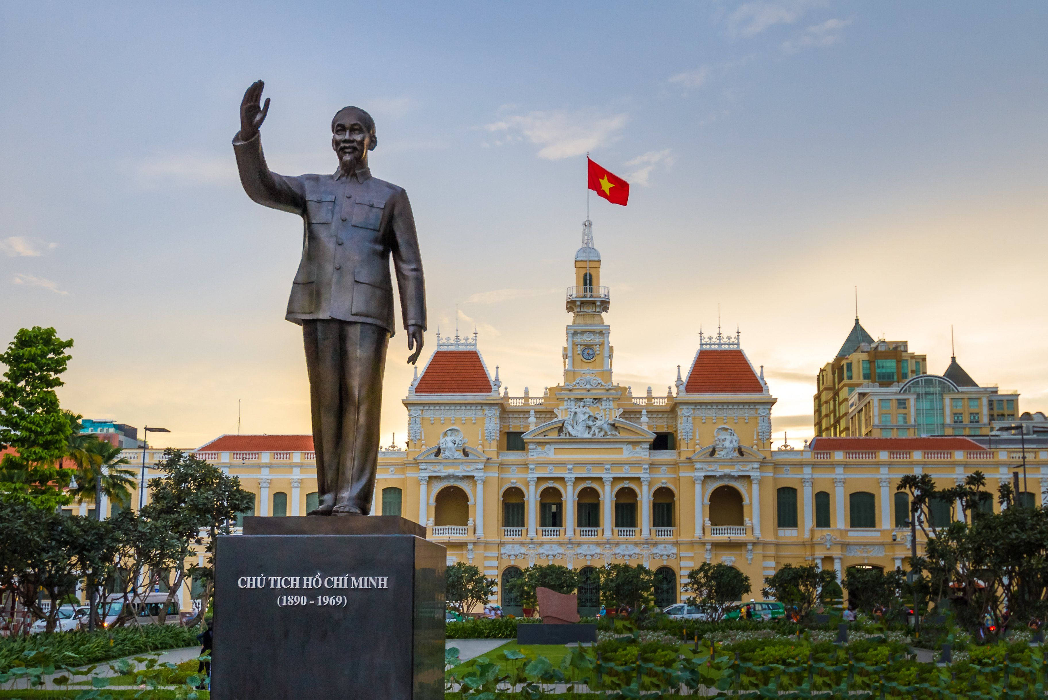 胡志明市銅像 壁紙 — 胡志明銅像喺日落時分喺一座宏偉嘅黃色殖民建築旁邊揮手，建築物上飄揚住一面紅旗。