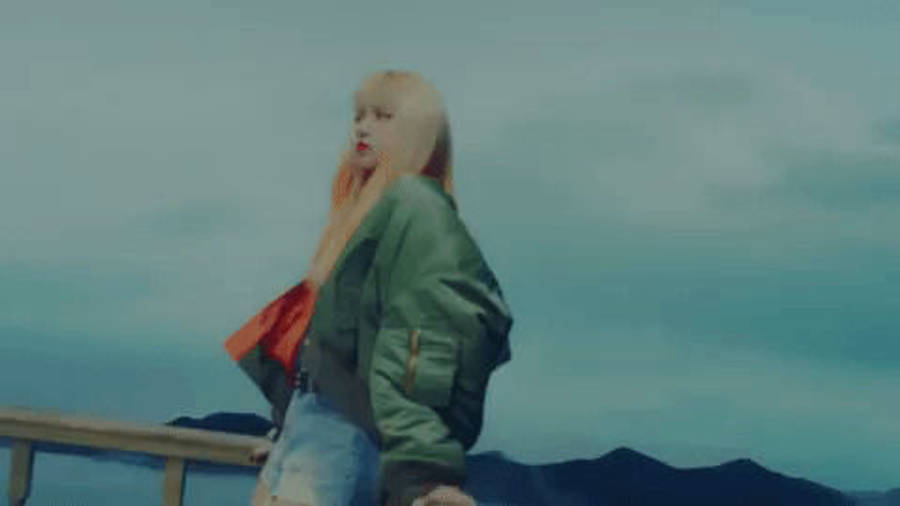 脈動舞后——BLACKPINK 的 Lisa 壁紙 — 一位金髮女子身穿綠色外套和牛仔褲，站在木製欄杆上，俯瞰多雲天空下的遠山。