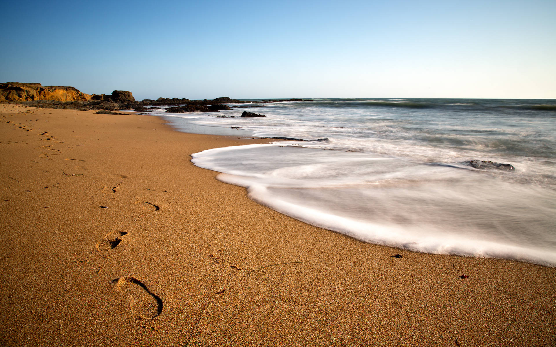 Beach Desktop 桌布 壁紙 — 腳印沿著金色沙灘延伸，溫柔的海浪在清澈的藍天下翻滾。