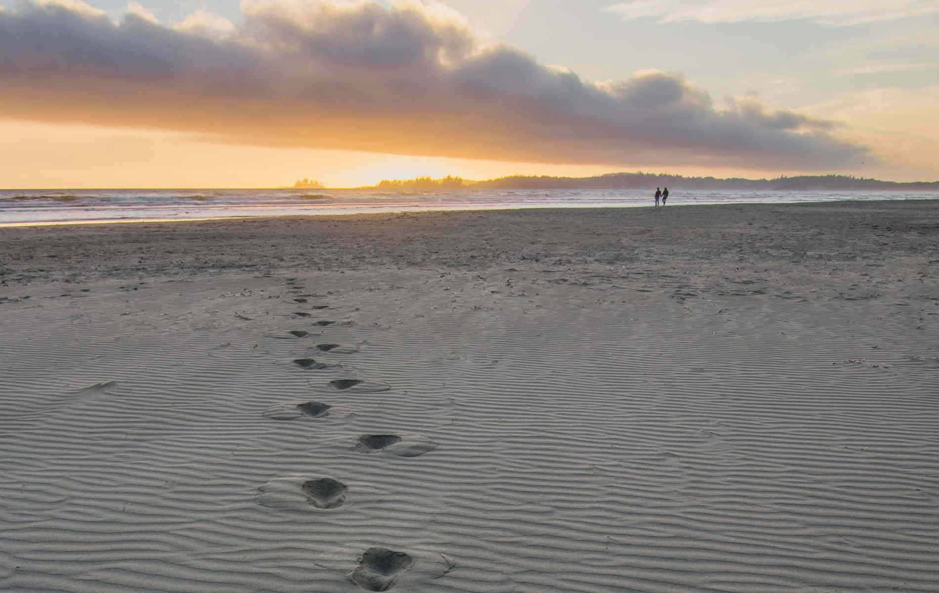 Beach Walk 桌布 壁紙 — 腳印穿過波紋狀的沙灘，朝向夕陽下廣闊海灘上的兩個遠處人影。