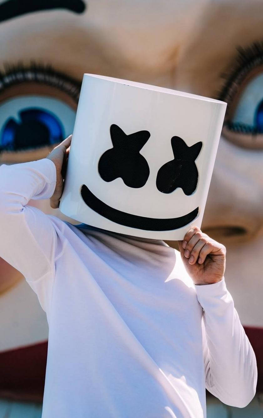 臉部背景Marshmello iPhone壁紙 壁紙 — 穿著白色襯衫的人手持標誌性的白色棉花糖面具，上面有黑色笑臉，背景是巨大的娃娃眼睛。
