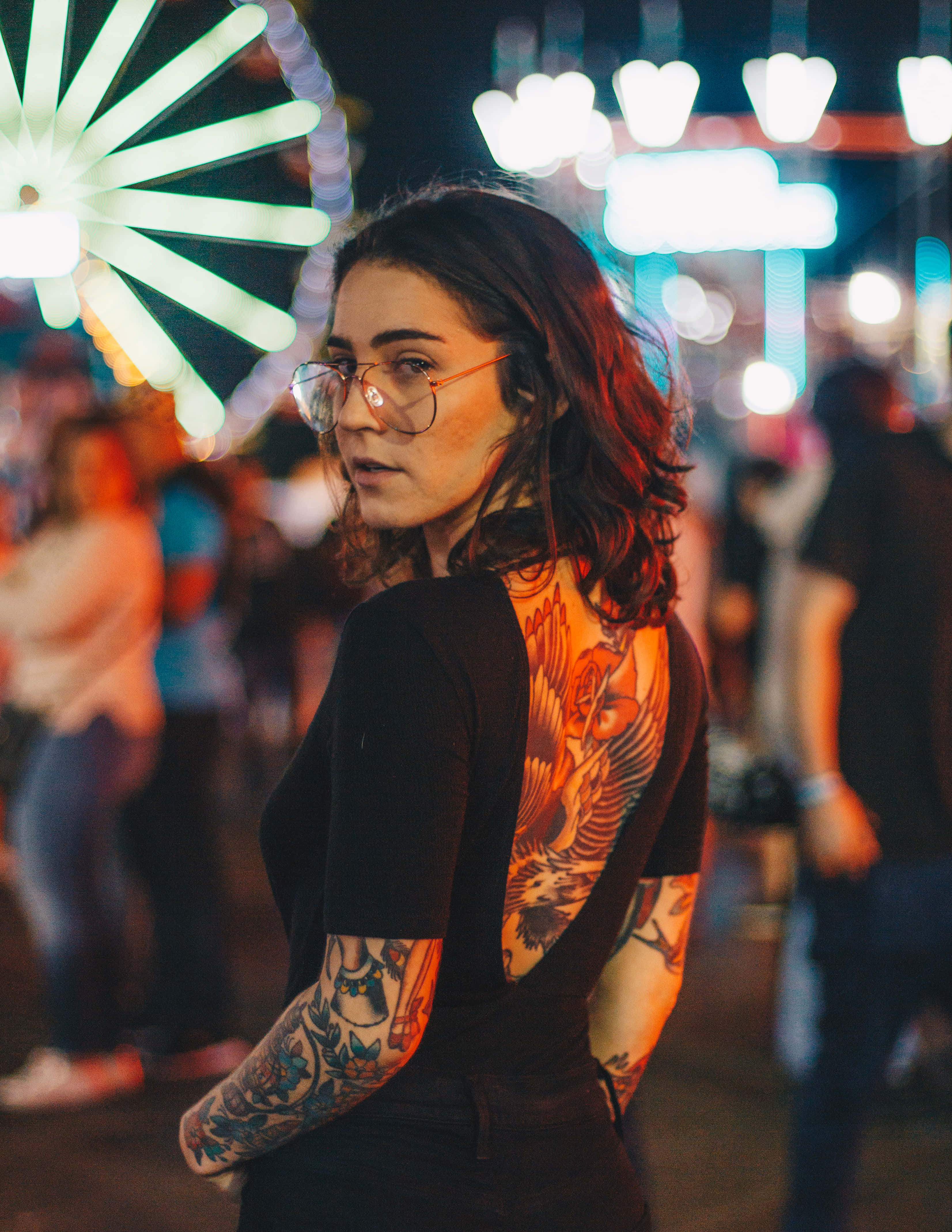 自信展現細緻紋身的女性 壁紙 — 一位戴眼鏡、擁有複雜紋身的女性在夜晚嘉年華的閃爍燈光下轉向鏡頭。