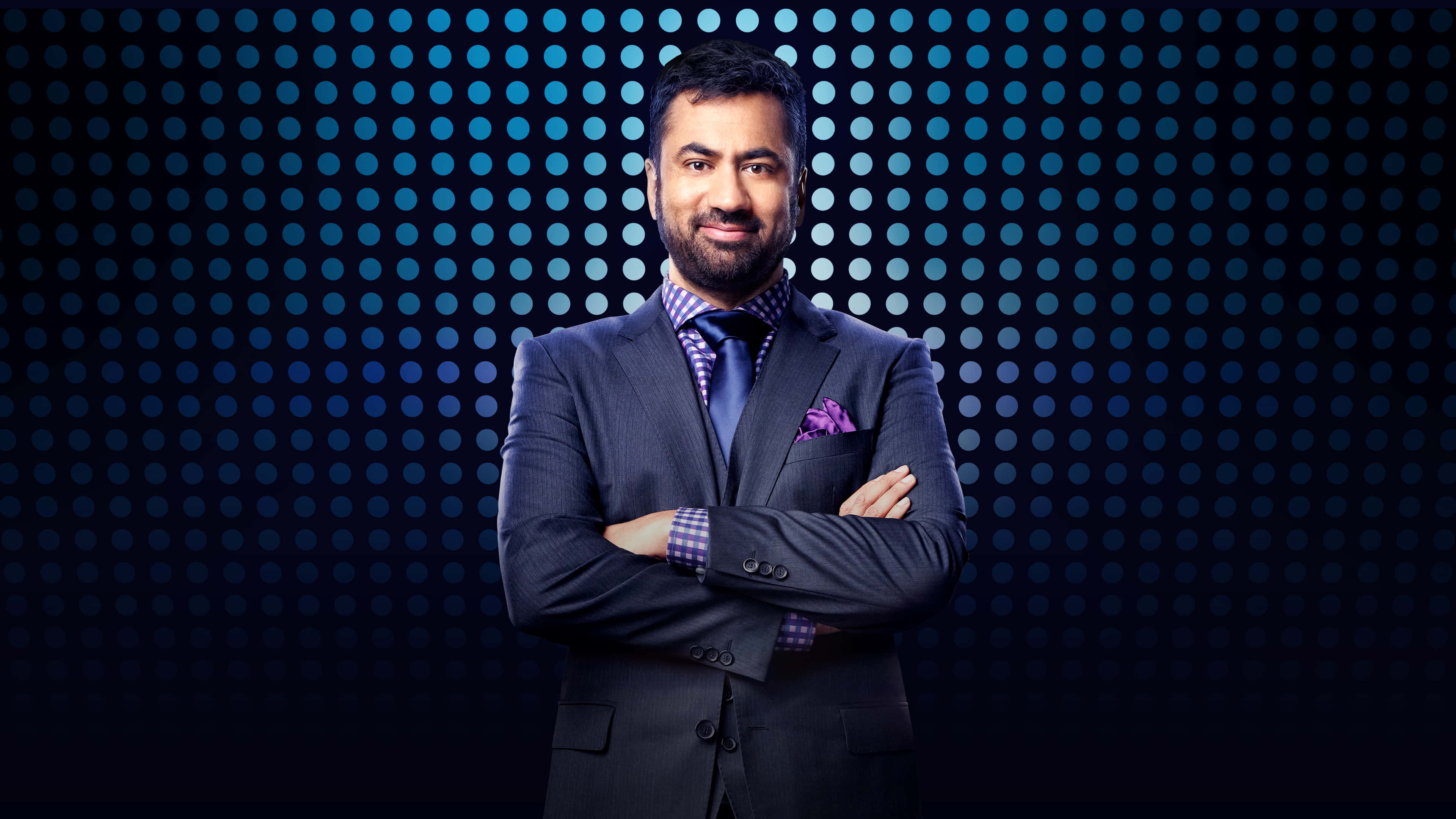 Kal Penn 桌布 壁紙 — 自信滿滿嘅男士著住剪裁俐落嘅西裝，雙手交叉企喺發光藍色圓點圖案背景前。