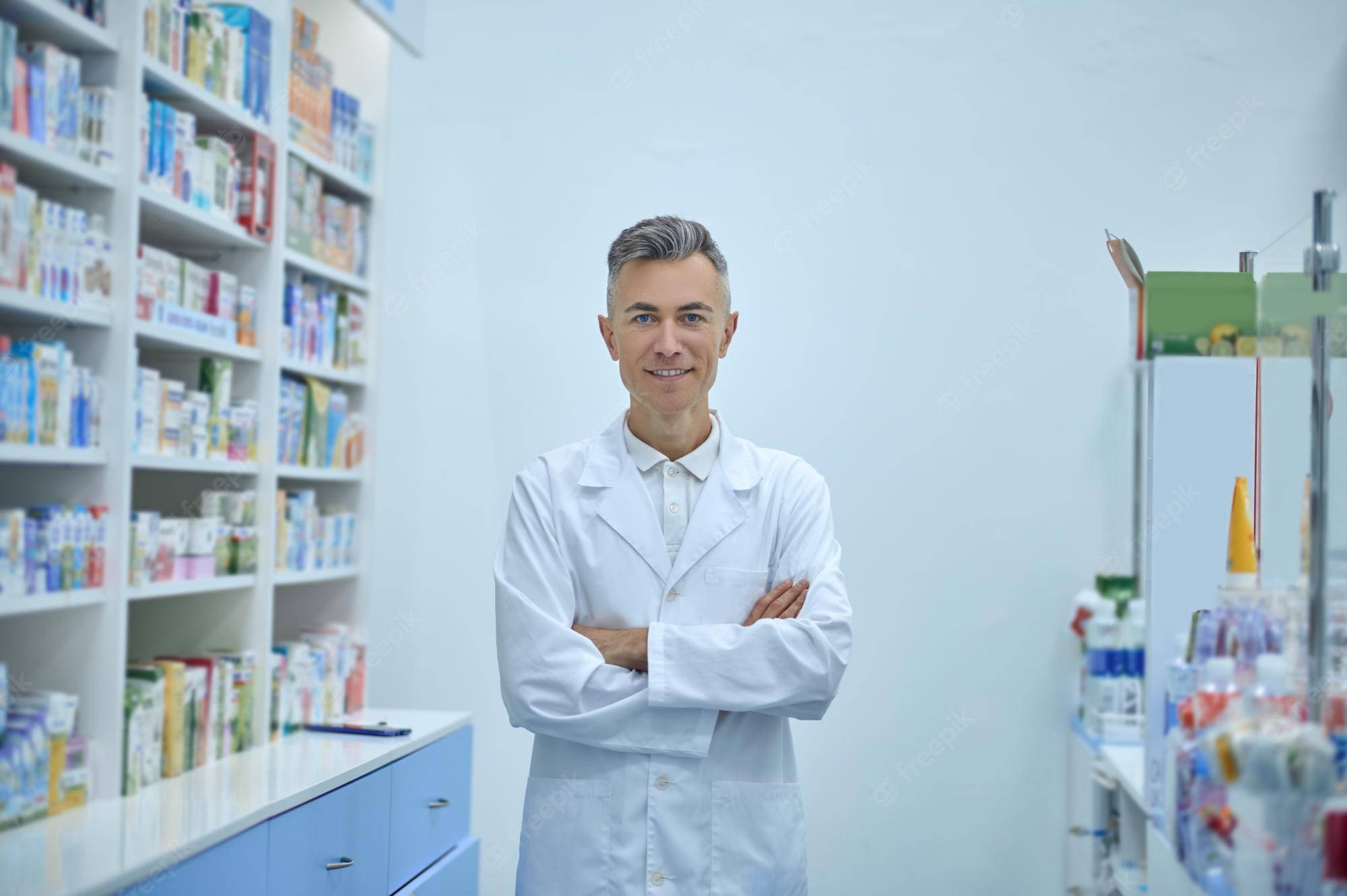 自信藥劑師雙臂交疊 壁紙 — 一位自信的藥劑師身穿白袍，在光線充足的藥局走道中交叉雙臂站立，周圍是色彩繽紛的藥品貨架。