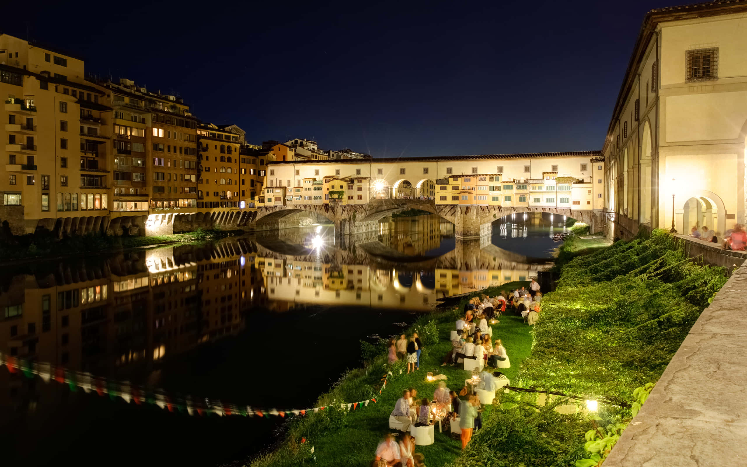 自助導覽舊橋 壁紙 — 夜晚亮燈的 Ponte Vecchio 橋，人們在溫暖燈光下的草河岸用餐。