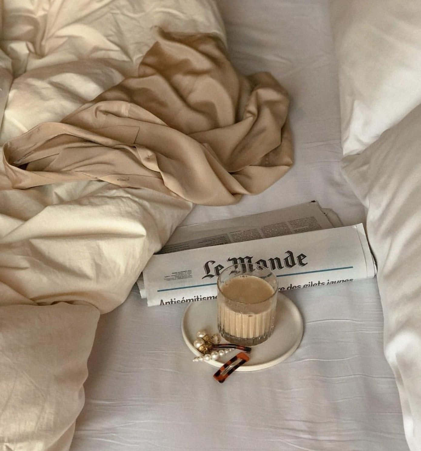 自然之美：棕色美學 壁紙 — 一張舒適的床，有皺褶的米色床單、報紙、一杯拿鐵咖啡和白色碟上的珍珠髮夾。