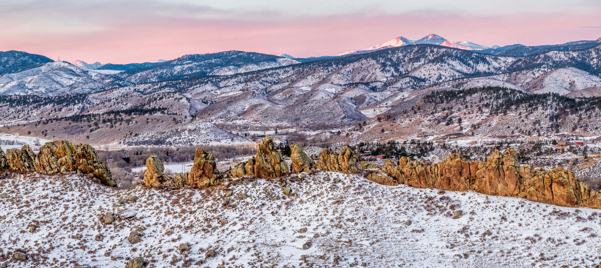 自然之美——科羅拉多山脈 壁紙 — 白雪覆蓋崎嶇的岩石地貌和起伏的丘陵，在柔和的粉紅色和橙色暮色天空下，遠處有積雪的山峰。