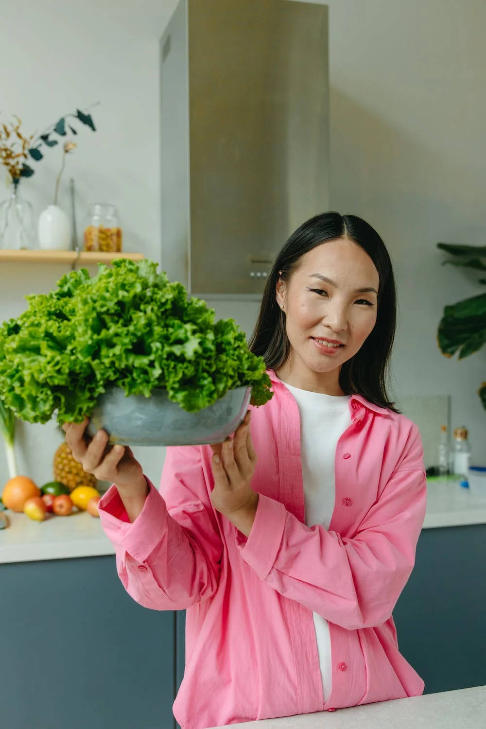自然光下新鮮生動綠色生菜 壁紙 — 一位身穿粉紅色襯衫的微笑女士，在現代廚房裡自豪地捧著一大碗新鮮的綠色生菜。