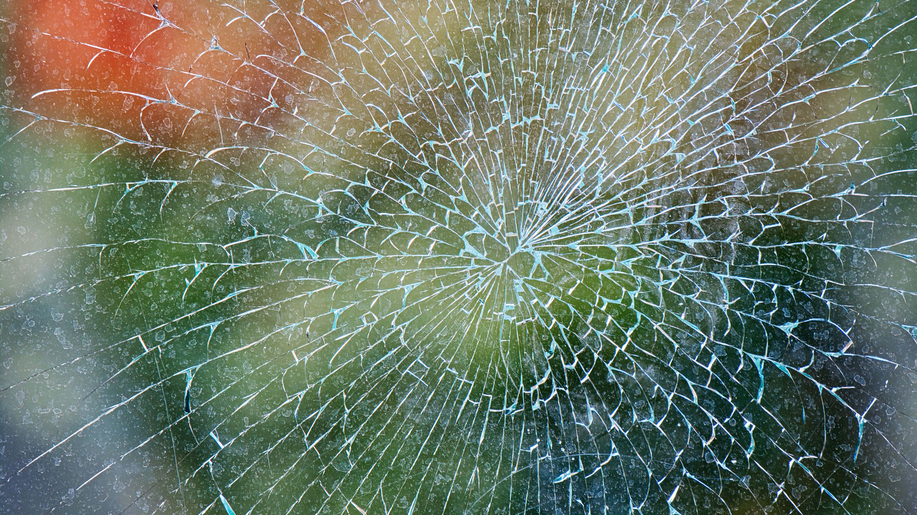 自然模糊破碎玻璃圖像 壁紙 — 碎裂玻璃的特寫，帶有放射狀裂紋圖案，顯露出模糊的綠色和橙色背景。