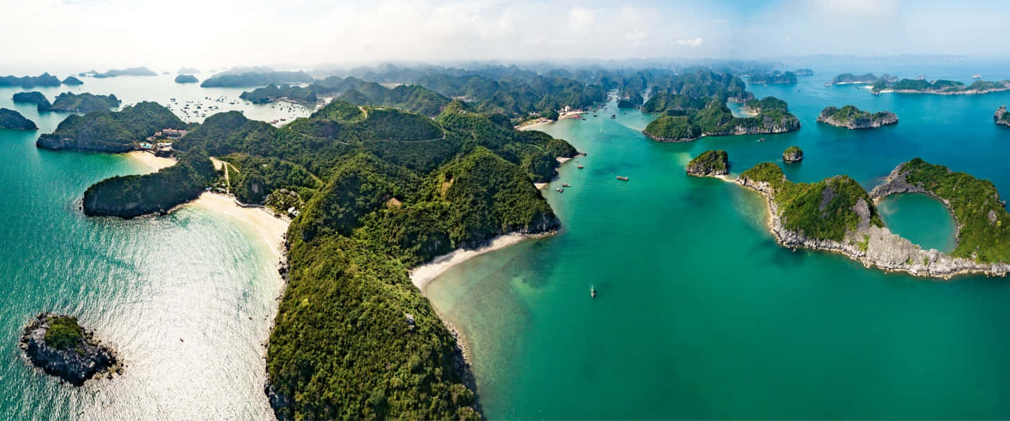 自然瀑布的壯麗景象 壁紙 — 鳥瞰翡翠綠的海水環抱著翠綠的喀斯特島嶼，沙灣和散落的船隻在明亮的天空下。