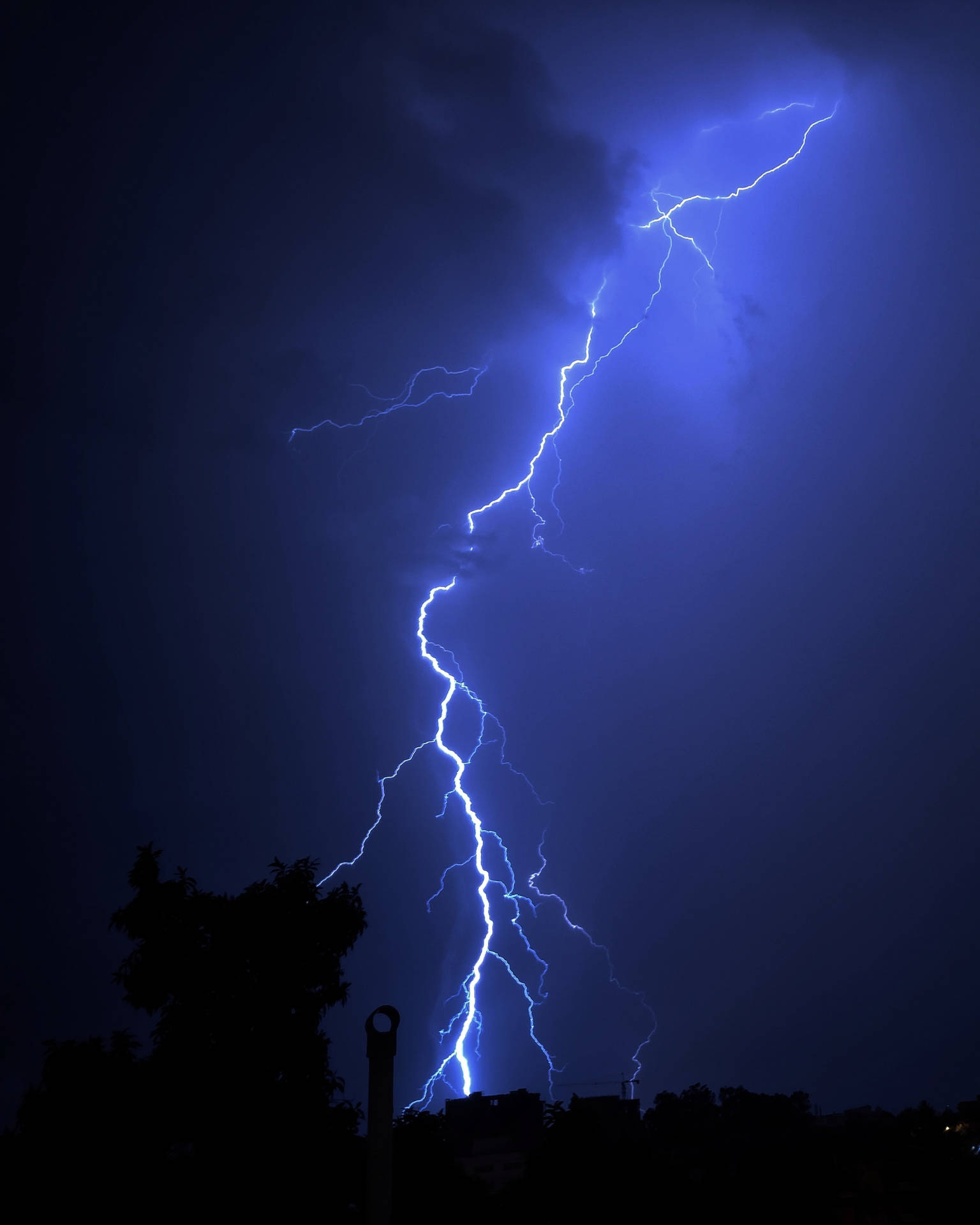 強大的藍色閃電照亮了暴風雨的夜空，映襯出黑暗的剪影樹木和建築物。